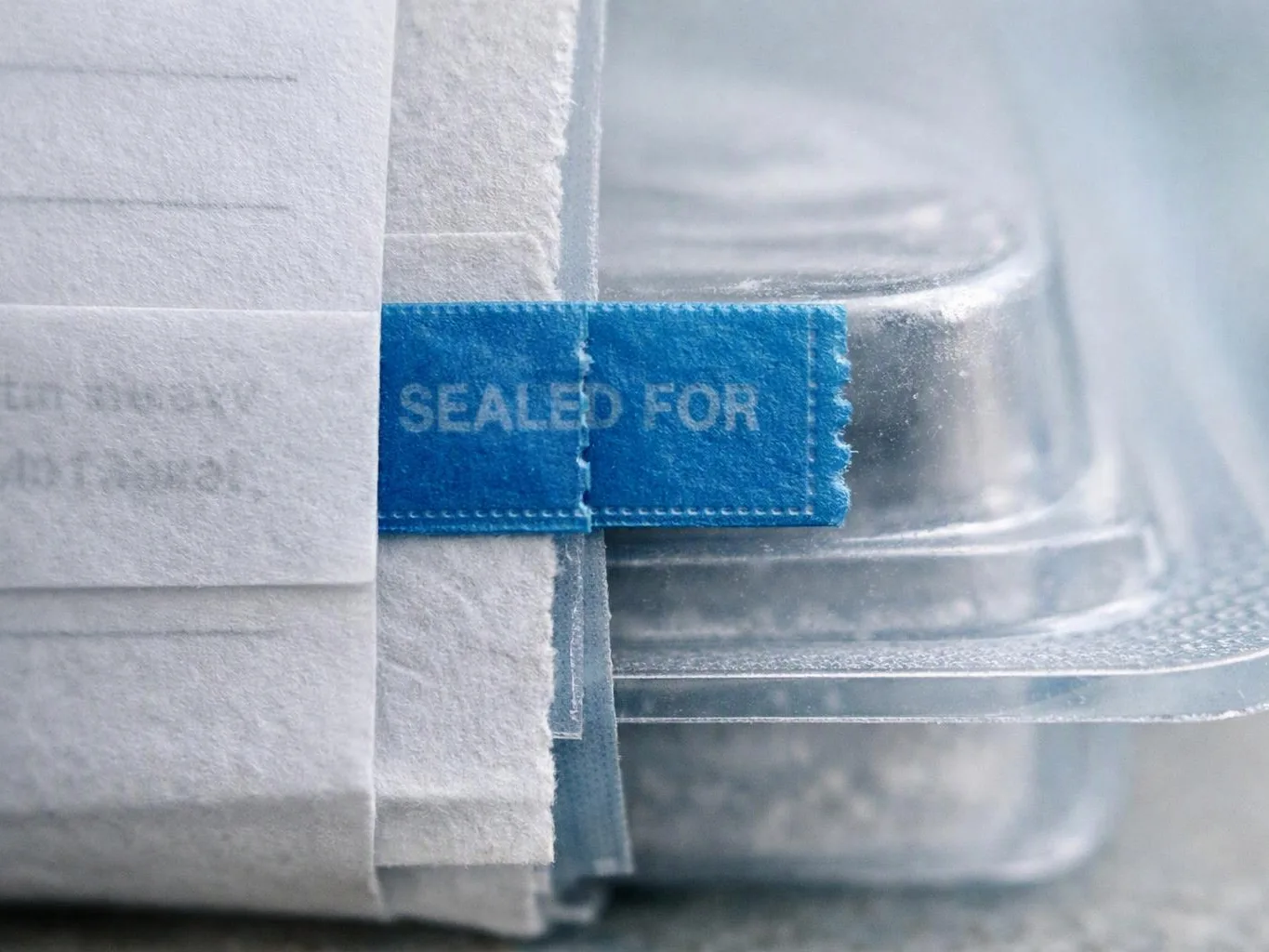 Close-up of a blue tamper seal on a wrapped clear plastic container with folded white paper beside it