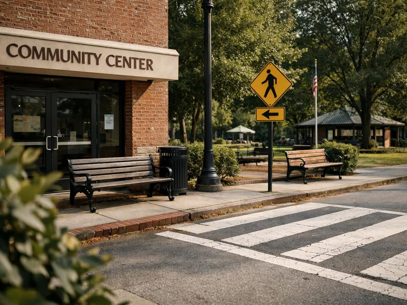 A community center entrance faces a crosswalk, with benches, a pedestrian sign, and trees in a quiet park-like setting