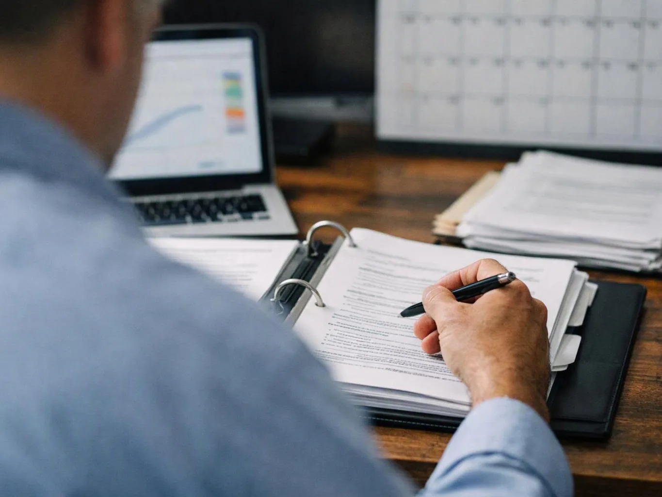 A person in a blue shirt reviews papers and writes in a binder beside a laptop and calendar on a desk