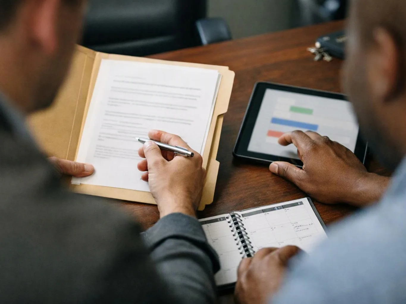 Two people review papers and a tablet with colored bars at a wooden table